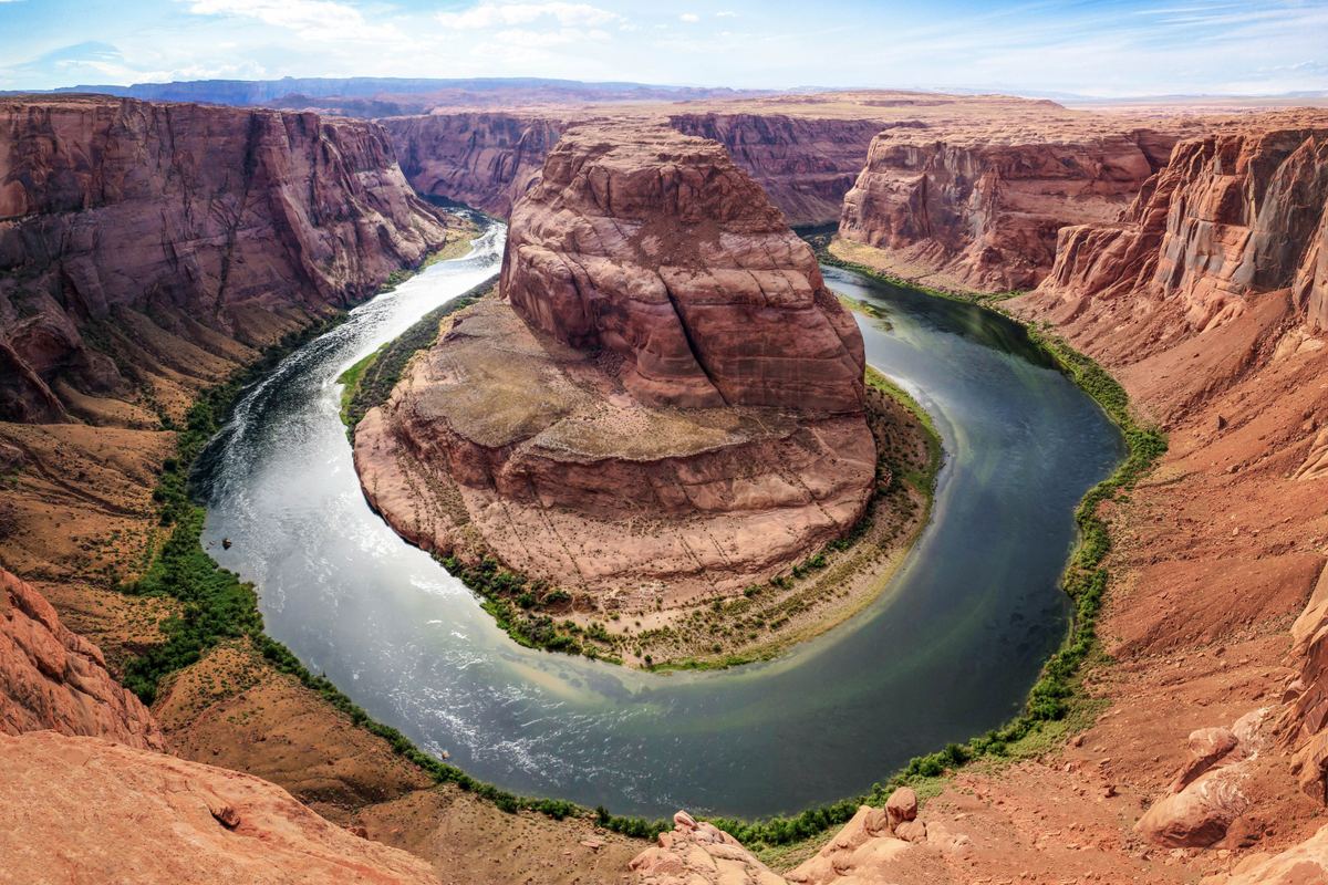 A river curves in a tight bend around a large red rock butte, with green vegetation along the banks and canyon walls rising on both sides.
