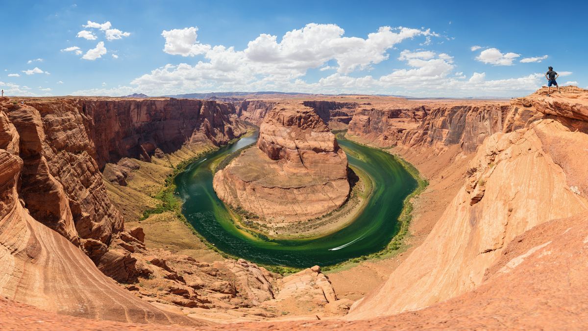 Wide panorama of a green river bending around a rock butte in a deep red sandstone canyon under a blue sky with white clouds.